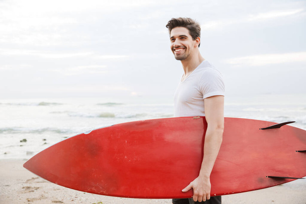 Handsome young man surfer with surfing on a beach outside. - Zdjęcie, obraz