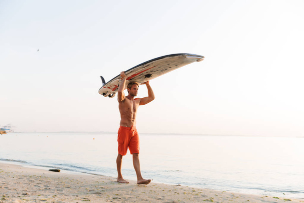 Image of muscular surfer man carrying his surfboard by ocean at  - Фото, зображення