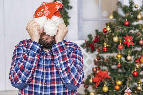περιμένοντας για ένα θαύμα έννοια Χριστούγεννα φορά γενειοφόρος άνθρωπος πορτρέτο κρύβει το πρόσωπό του πίσω από τον Άγιο Βασίλη καπέλο και τα χέρια, εορταστική λευκό εσωτερικό με διακοσμημένο δέντρο  - Φωτογραφία, εικόνα
