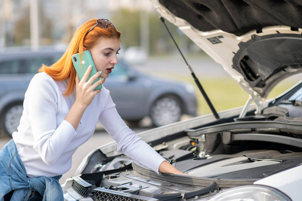 Young woman standing near broken car with popped hood talking on - Foto, imagen