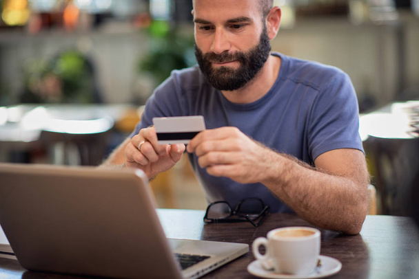 Guapo barbudo caucásico hombre sentado en la cafetería, la celebración de la tarjeta de crédito y el uso de ordenador portátil para las compras en línea
. - Foto, imagen