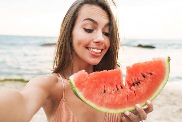 Image of amusing woman eating watermelon and taking selfie photo - 写真・画像