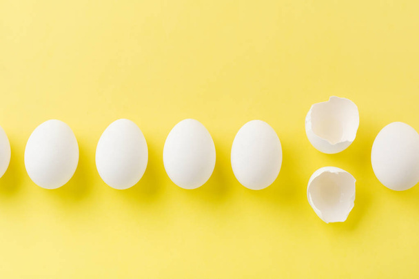 White raw chicken eggs lying in horizontal row with broken egg on yellow background. Top view. - 写真・画像