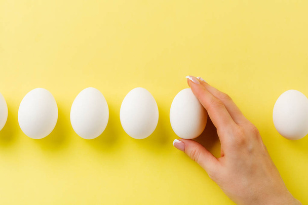 White raw chicken eggs lying on yellow light background and female hand holds broken egg. Top view. - Foto, Bild