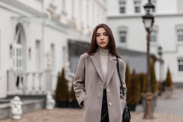 黒のスカートの中に革のハンドバッグを持つエレガントなコートの現代的なかわいい若い女性は、ヴィンテージの白い建物の近くの通りに沿って歩いています。秋の日に街の流行の服でヨーロッパの都市の女の子. - 写真・画像