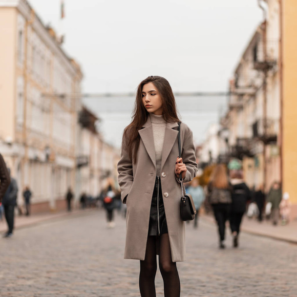 黒のスカートの中に革のハンドバッグを持つエレガントなコートのかわいい魅力的な若い女性は、ヴィンテージの建物の近くの通りに沿って歩いています。秋の日に街の流行の服で美しい都市の女の子. - 写真・画像