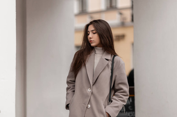 Moderní městská mladá žena model v elegantním kabátě s koženou kabelkou stojí na ulici v blízkosti vinobraní bílé budovy. Evropská městská dívka v módním oblečení ve městě v podzimním dni. - Fotografie, Obrázek