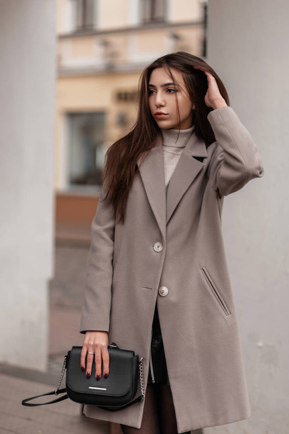 Pretty young brunette woman in a fashionable long coat with a stylish leather black handbag poses in the city near a white vintage building. Modern cute girl outdoors.Trendy seasonal women's outerwear - 写真・画像