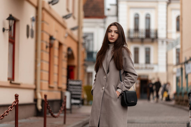 Vonzó fiatal nő hosszú, fényűző barna hajjal, elegáns kabátban, divatos fekete bőrtáska áll a városban, közel a vintage épületekhez. Gyönyörű lány modell pózol a szabadban. - Fotó, kép