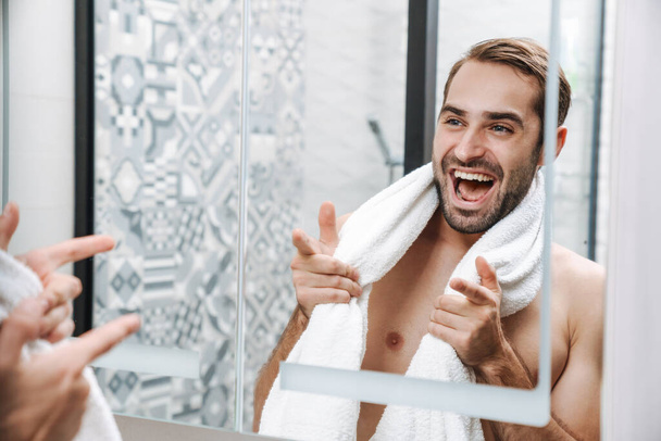 Happy young shirtless man looking at himself at the bathroom - 写真・画像