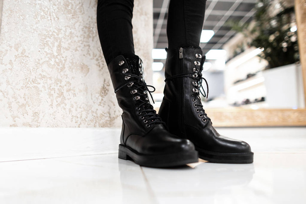 Stylowa młoda kobieta stojąca na płytce w sklepie w czarnych, fajnych, skórzanych butach z klasycznymi dżinsami. Zamknij kobiece nogi w modnych jesienno-wiosennych butach. Obuwie z kolekcji sezonowej. - Zdjęcie, obraz
