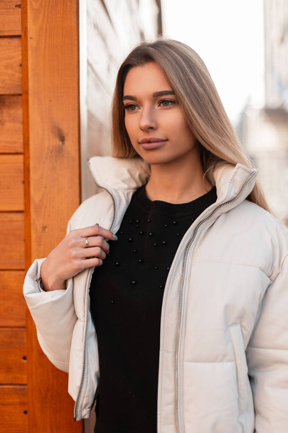 Retrato de una hermosa joven con el pelo rubio en una chaqueta blanca de moda en un suéter negro vintage en la calle cerca de un edificio de madera. Atractiva chica moderna con maquillaje natural en la ciudad
. - Foto, Imagen