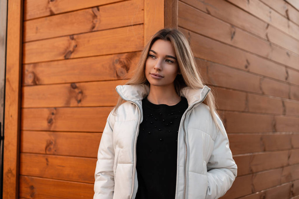 European pretty young woman blonde in a black stylish sweater in a stylish white jacket stands on the street near a wooden vintage building. Attractive girl fashion model on a warm autumn day outdoors - Photo, Image