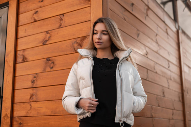Aranyos fiatal európai nő szőke egy vintage fehér tavaszi kabát egy elegáns fekete pulóver pózol közelében egy barna fa épület az utcán. Divatos csinos városi lány divat modell szabadban. - Fotó, kép