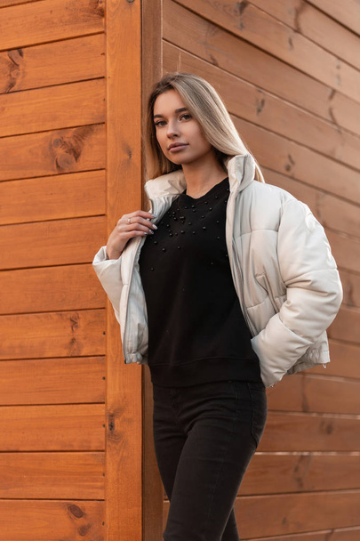 Young attractive blonde woman in a black sweater in a fashionable white jacket stands on the street near a brown wooden building. Cute girl in trendy clothes is enjoying a walk in the city. Style. - Photo, Image