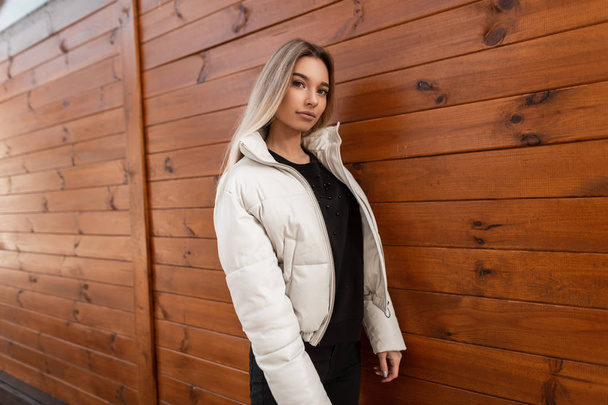European glamorous young woman blonde in a fashionable white jacket in black jeans in a sweater stands outdoors near a wooden vintage wall. Attractive modern girl fashion model walks on the city. - Foto, imagen