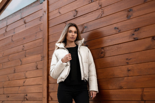 Glamorous young woman blonde with sexy lips in a black stylish sweater in a fashionable white winter jacket posing outdoors near a wooden vintage building. Cute girl standing in city. Youth fashion. - Photo, Image
