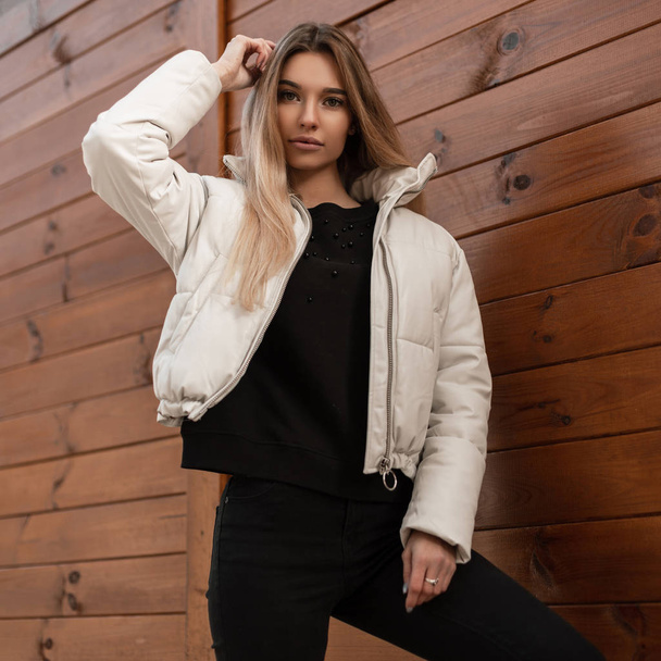 Pretty trendy young woman model in a stylish white jacket in fashionable black clothes posing outdoors near a vintage wooden wall. Beautiful blonde girl in the street. Fashion youth women's clothing. - Foto, immagini