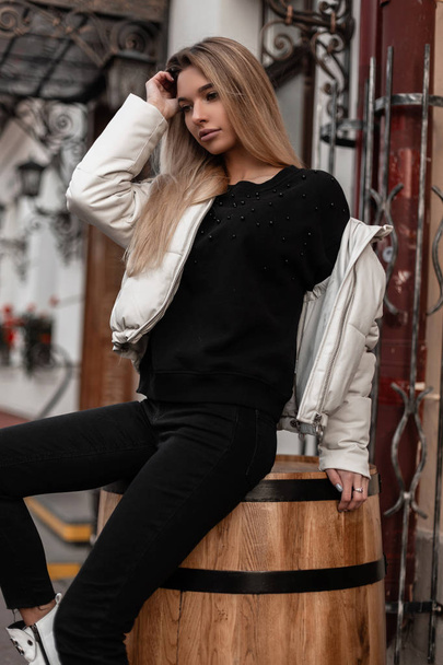 European young blonde woman in a fashionable white jacket in black stylish jeans in a knitted vintage sweater is resting sitting on a wooden barrel in the city. Trendy girl outdoors on a autumn day. - Photo, Image