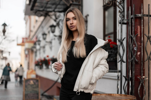 Model uroczej młodej kobiety w modnej białej kurtce w czarnym swetrze na świeżym powietrzu w mieście, w pobliżu zabytkowego budynku. Całkiem stylowa dziewczyna chodzi po ulicy w modnych ubraniach. Styl wiosenny. - Zdjęcie, obraz
