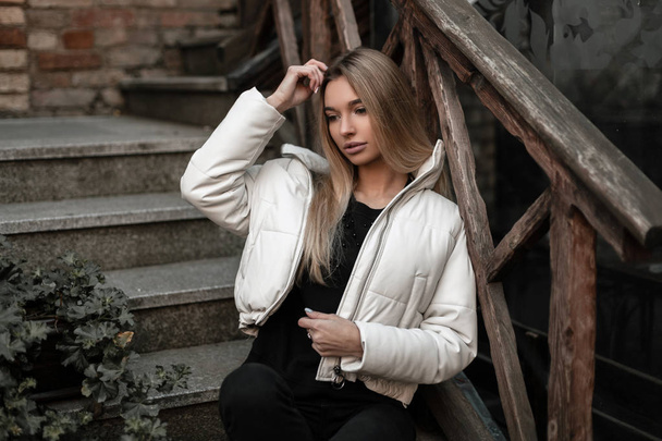 Trendy young woman in fashionable clothes with long blond hair is resting in the city on a stone staircase with vintage wooden railing. Beautiful girl model in a stylish outerwear outdoors. Autumn. - Foto, immagini