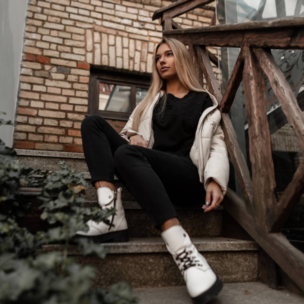 Mujer joven europea glamorosa rubia en ropa de temporada de moda sentada en una escalera de piedra. Linda chica en una chaqueta blanca de moda en vaqueros negros en botas de cuero con estilo está descansando al aire libre
. - Foto, Imagen