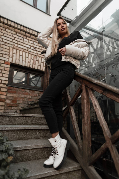 Urban-Fashion-Modell einer niedlichen jungen Frau in Frühlings- und Herbstkleidung in trendigen Stiefeln, die in der Stadt auf einer Treppe mit altem Holzgeländer posiert. Attraktives Mädchen in stylischer Oberbekleidung steht draußen. - Foto, Bild