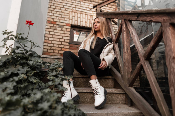 Pretty stylish young woman model in a fashionable white jacket in black jeans in trendy autumn white leather boots is resting on a stone staircase with wooden railing. Beautiful cute blonde girl. - Photo, Image