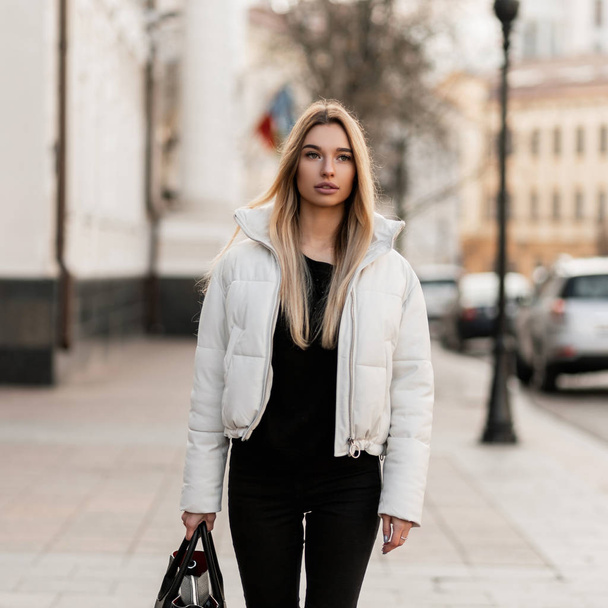 Trendy mladá blondýnka v černobílém módním oblečení s koženou stylovou taškou kráčí po ulici za teplého podzimního dne. European girl fashion model enjoys a walk in the city. - Fotografie, Obrázek