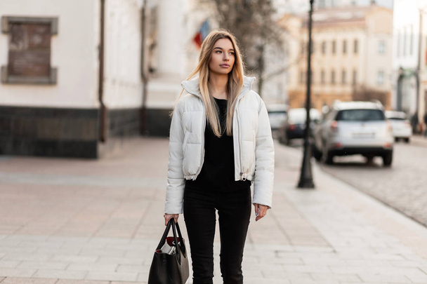 Krásná mladá žena s blond vlasy v módní bílé bundě v džínách s černou stylovou koženou taškou na ulici. Atraktivní módní dívka model cestuje po městě. Styl jaro-podzim. - Fotografie, Obrázek