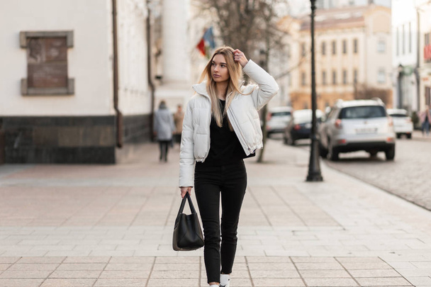 Bella giovane donna urbana in una giacca bianca alla moda in jeans neri vintage con una borsa in pelle nera elegante per strada. Attraente modello ragazza di moda viaggia in giro per la città. Stile autunno
. - Foto, immagini