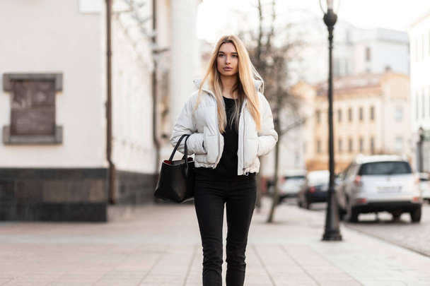 Modelo de mulher muito jovem magro com cabelo loiro longo em uma jaqueta branca na moda em jeans pretos elegantes em uma bolsa de couro na moda caminha ao longo de uma rua da cidade em um dia quente de outono. Menina moderna está andando
. - Foto, Imagem