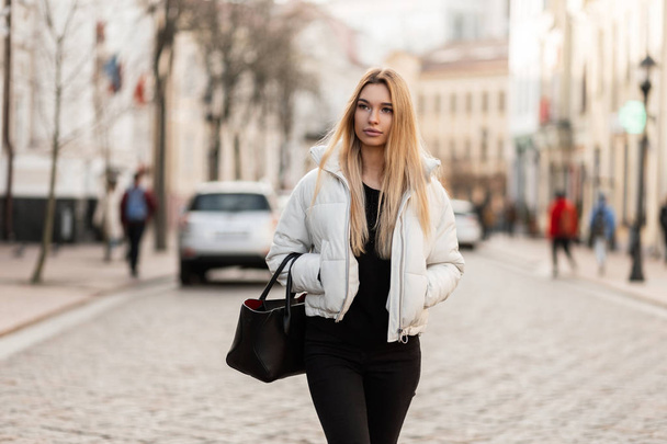 流行のレザーバッグにブラックジーンズでファッショナブルな白い季節のジャケットに長いブロンドの髪を持つ若い女性モデルは、暖かい秋の日に街の通りに沿って歩いています。かわいい都会の女の子が歩いている。様式. - 写真・画像