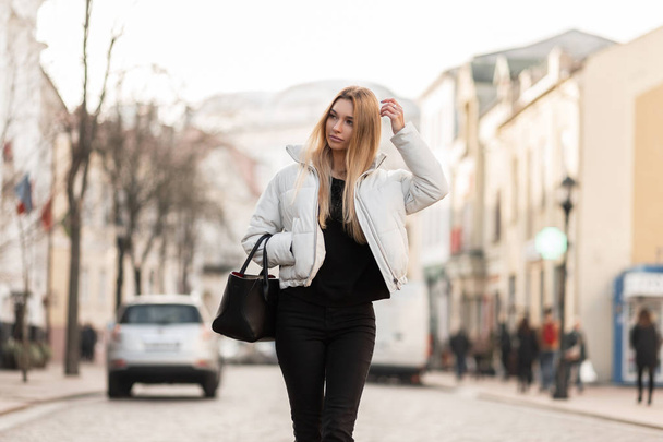 魅力的な若いブロンドの女性は、革のハンドバッグとヴィンテージブラックジーンズのスタイリッシュな白いジャケットで街を歩いています。プリティービューティガールモデルが暖かい春の日に通りを歩く. - 写真・画像
