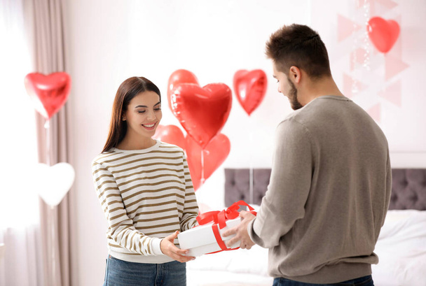 Young man presenting gift to his girlfriend in bedroom decorated with heart shaped balloons. Valentine's day celebration - Photo, Image