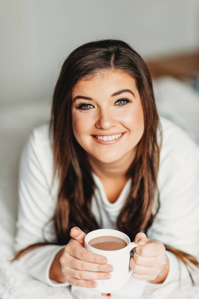 Retrato interior da jovem mulher feliz relaxando no quarto, vestindo pijama, segurando xícara de chocolate quente
 - Foto, Imagem