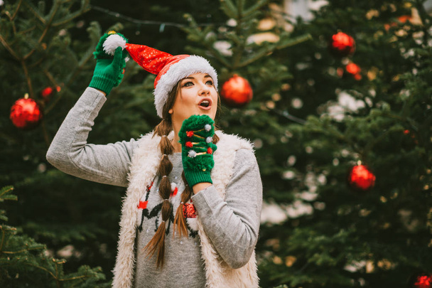 Festive portrait of happy young woman posing outside by the Christmas tree, wearing green mittens - 写真・画像
