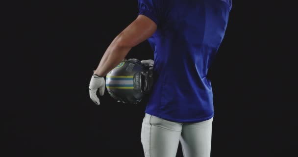 Rear view mid section of a Caucasian male American football player wearing a team uniform with pads, standing with his hand on his hip, holding a helmet and putting it under his arm, slow motion - Filmagem, Vídeo