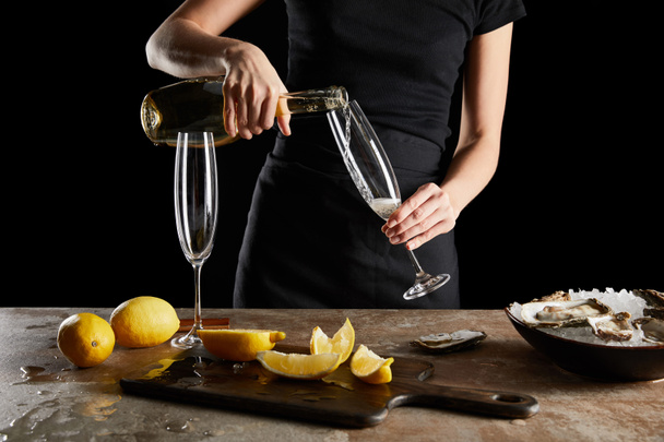 cropped view of woman pouring sparkling wine in champagne glass near oysters and lemons isolated on black  - Photo, Image