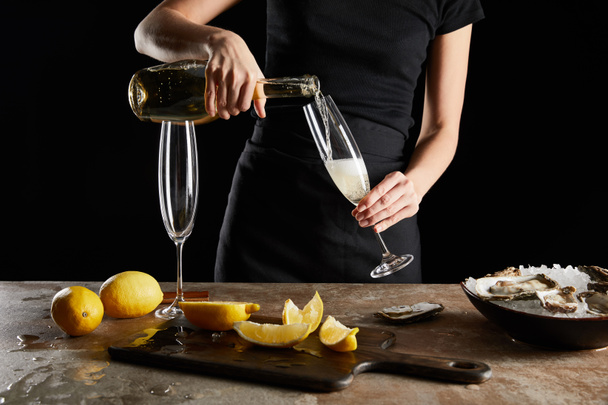 レモンの近くのシャンパングラスにスパークリングワインを注ぐ女性と黒で隔離されたカキのクロップドビュー  - 写真・画像