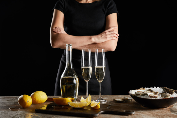 обрізаний вид жінки, що стоїть з схрещеними руками біля ігристого вина в пляшці та устриці в мисці з льодом ізольовані на чорному
  - Фото, зображення