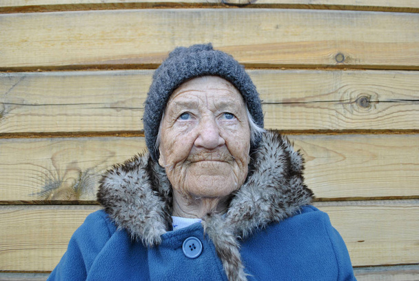 Uma avó idosa olha para cima. Vestida com um casaco quente com pele, e em sua cabeça um chapéu. No fundo é um fundo de madeira
. - Foto, Imagem