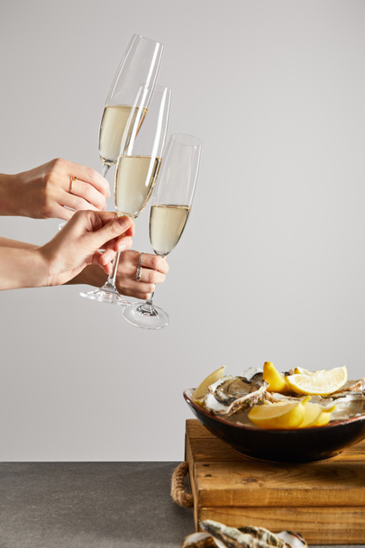 przycięty widok kobiet brzęczących kieliszkami szampana z winem musującym w pobliżu miski z ostrygami i świeżymi cytrynami izolowanymi na szarości  - Zdjęcie, obraz