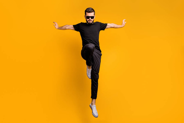 Full body photo of crazy guy jumping high practicing self defense kicking confident facial expression wear sun specs black t-shirt pants isolated yellow color background - Foto, Bild