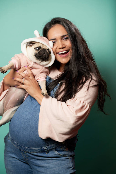 Pregnant brunette woman with pug in her hands on empty blue background - Foto, Imagen