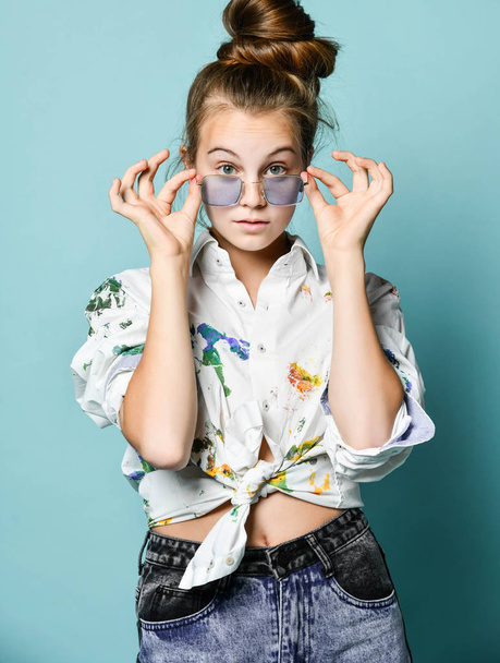 Portrait of surprised teenager girl in white shirt with paint stains tied at her stomach looking at us over glasses - Foto, Imagem