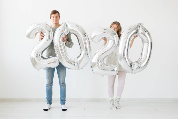 Γιορτές, εορταστική και κόμμα έννοια - Ευτυχισμένο ζευγάρι φιλιά και κρατά ασημένια μπαλόνια 2020 σε λευκό φόντο. Πρωτοχρονιά εορτασμού - Φωτογραφία, εικόνα