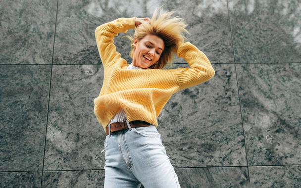 Pretty young woman wearing yellow sweater, blue hight waist jeans, jumping high, feeling happy on the city street. Student female dancing on the street, has joyful expression - Photo, Image