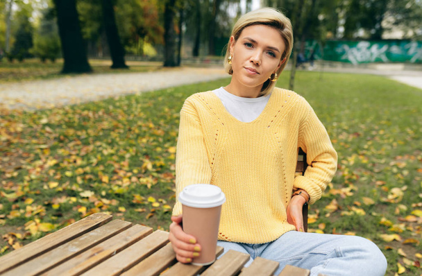 Beautiful woman drinking coffee at terrace cafe in the park. Portrait of young woman in the city street drinking hot cappuccino, looking at camera. Pretty woman with cup of coffee. Coffee on the go. - Foto, imagen