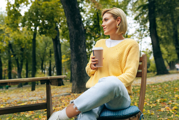 Beeld van een mooie jonge vrouw die koffie drinkt bij terrascafetaria in het park. Een vrouw in de straat die hete cappuccino drinkt en naar de camera kijkt. Mooi meisje met een kop koffie. Koffie onderweg.. - Foto, afbeelding
